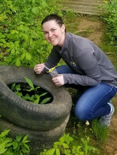 Photo of Dr. Parker, kneeling on a hiking trail, in a grassy area, next to a stack of auto tires.